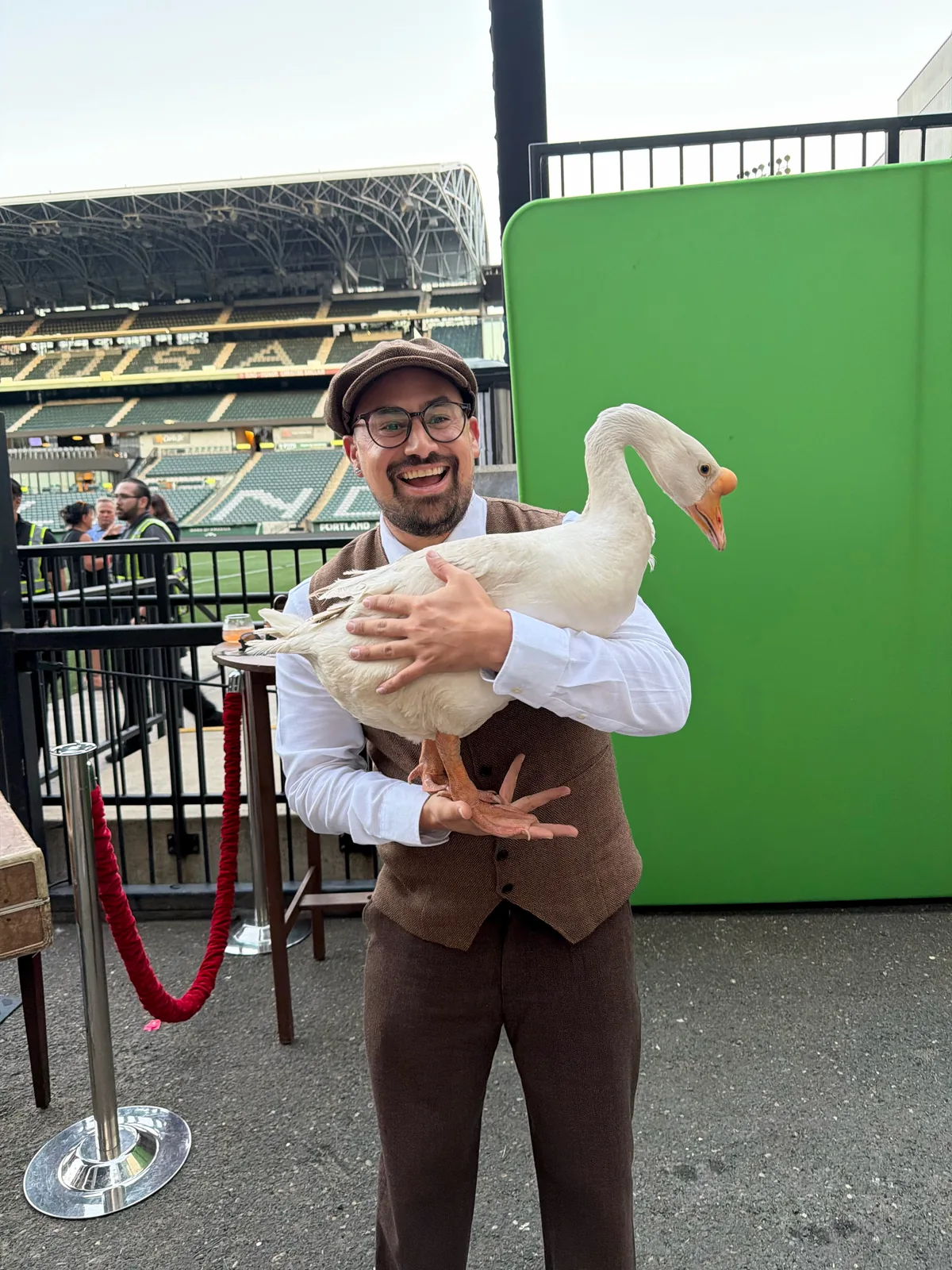 A smiling guest in a flat cap, glasses and brown vest holds Stella, a white goose, in front of a green screen, with the Providence Park stands behind him.
