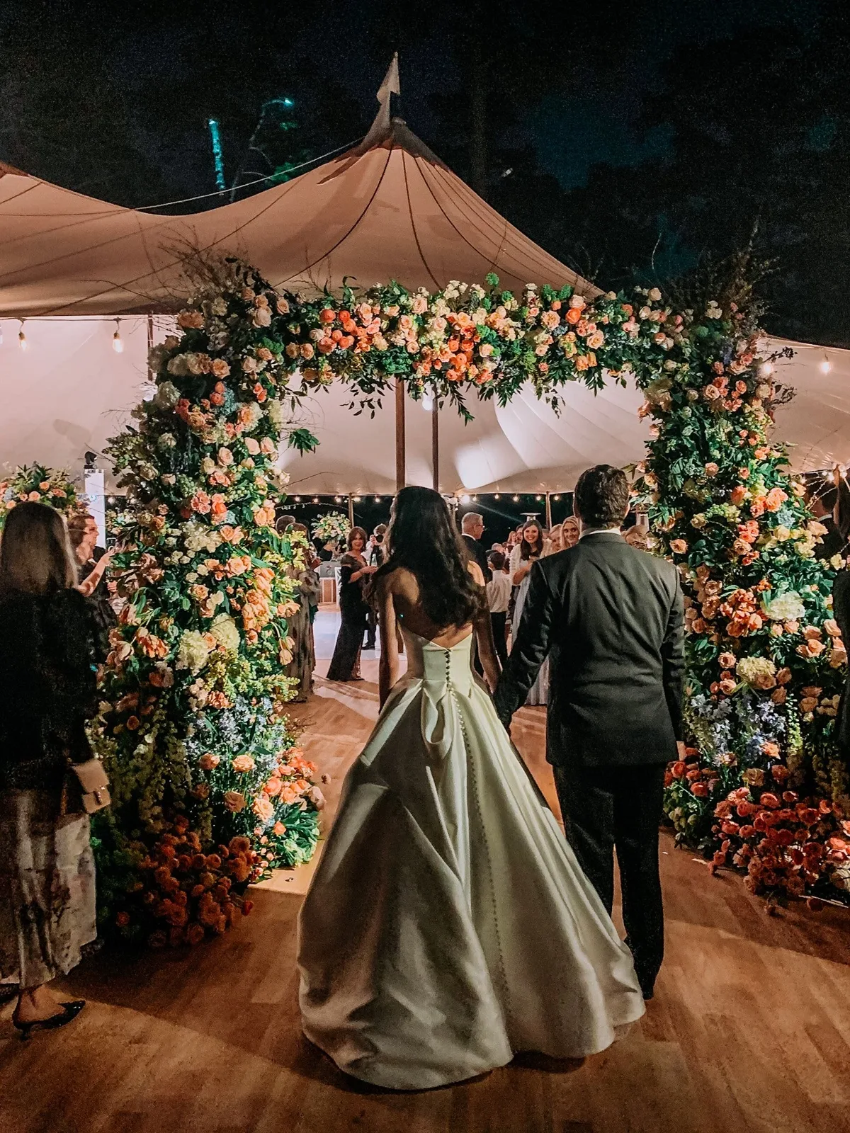Bride and groom walking into a tented reception under a floral arch