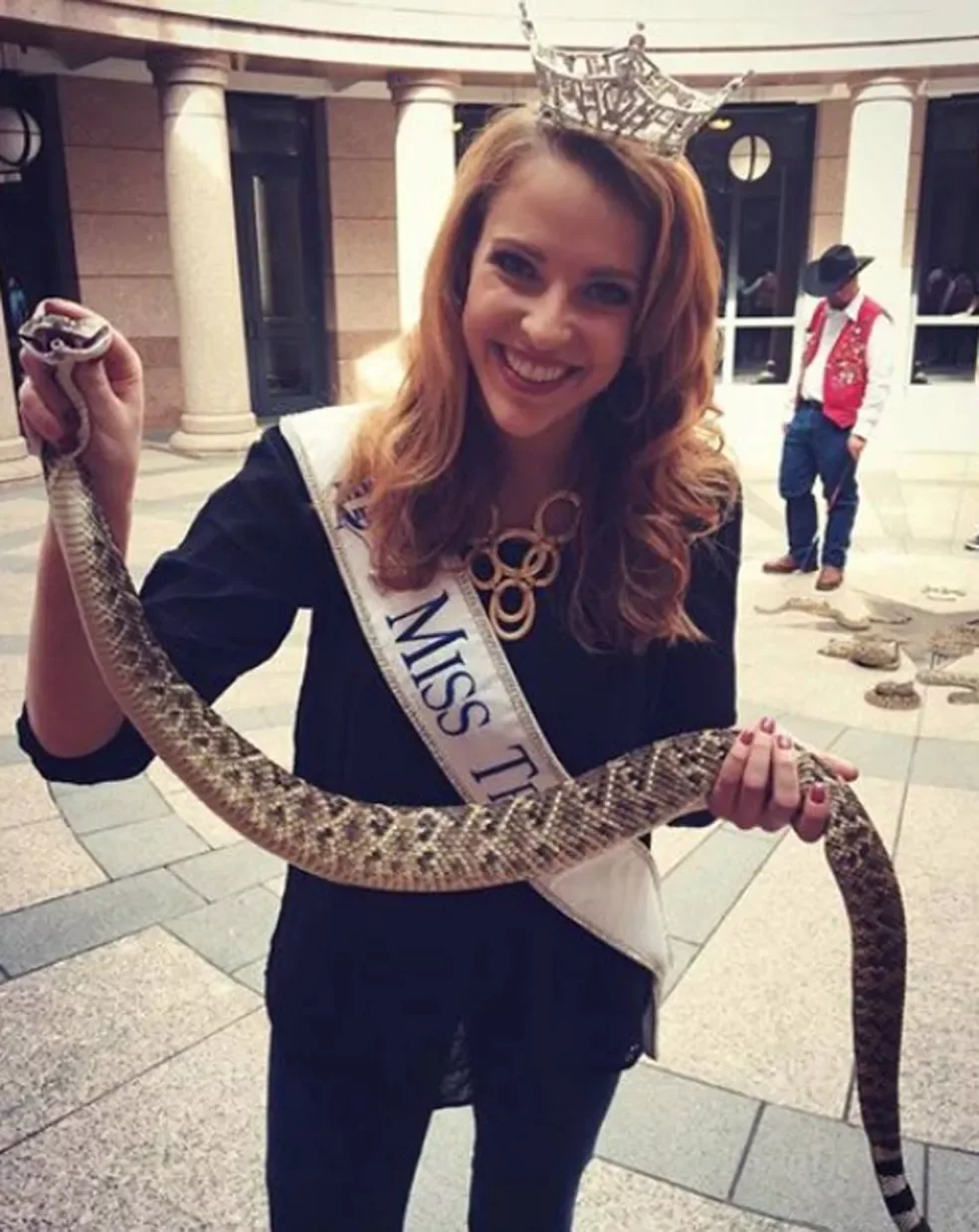 Miss Texas, wearing a crown and sash, smiles while holding a long rattlesnake with both hands. A man in a cowboy hat and red vest stands in the background near more snakes on the ground.