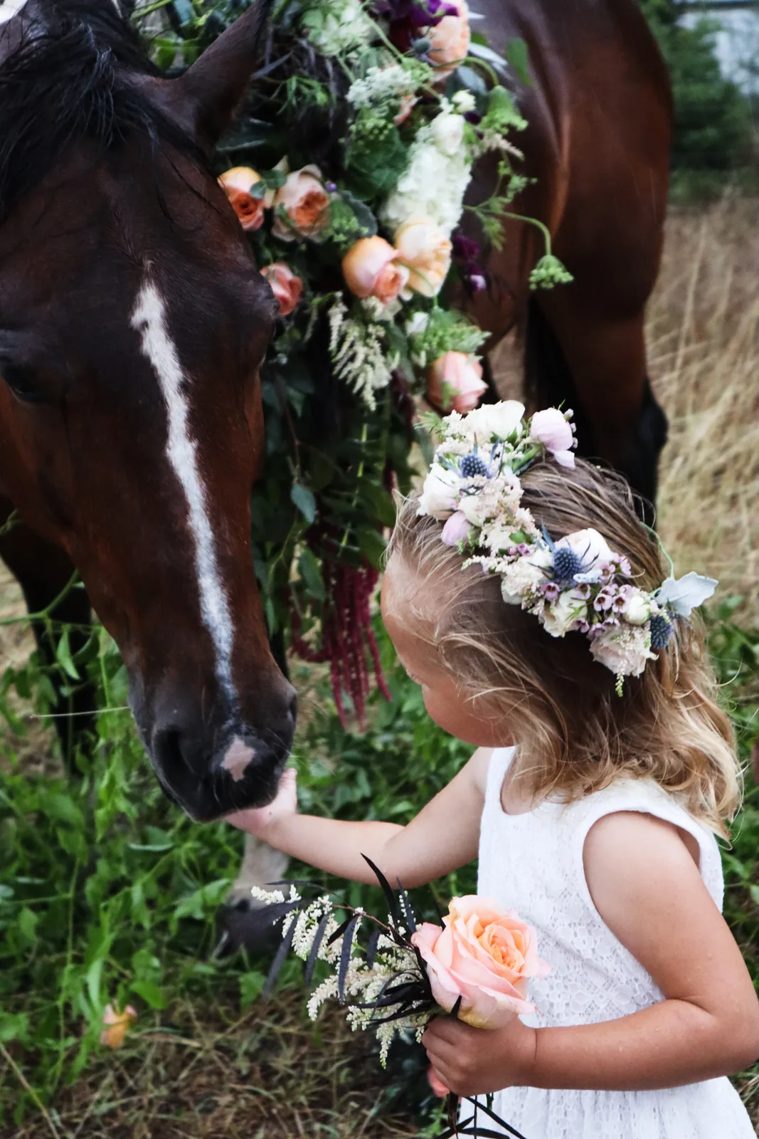 A young guest in a white dress and flower crown gently touches the nose of a brown horse wearing a large floral garland, holding a small bouquet in her other hand.