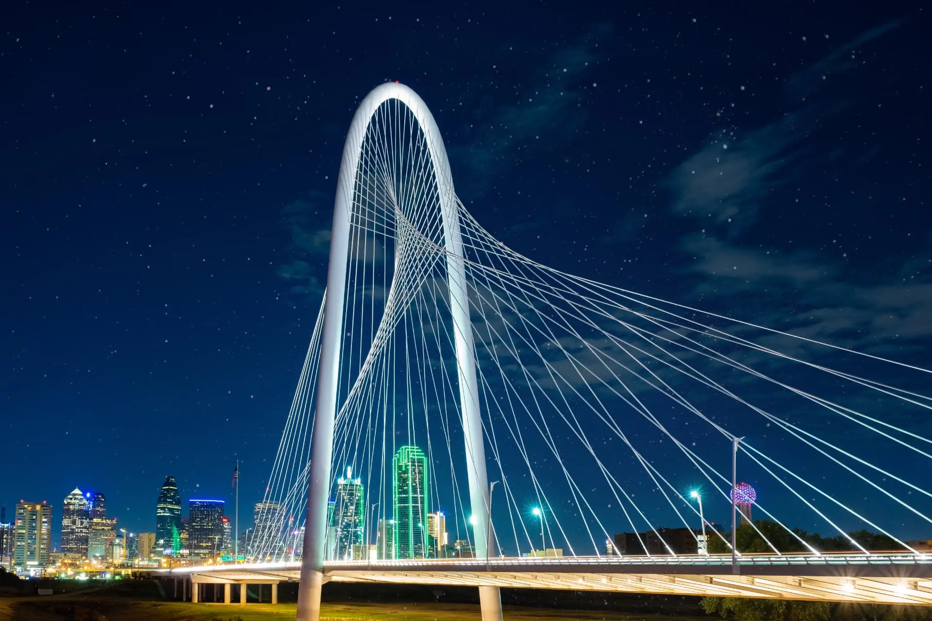 The Margaret Hunt Hill Bridge lit at night with the Dallas skyline behind it