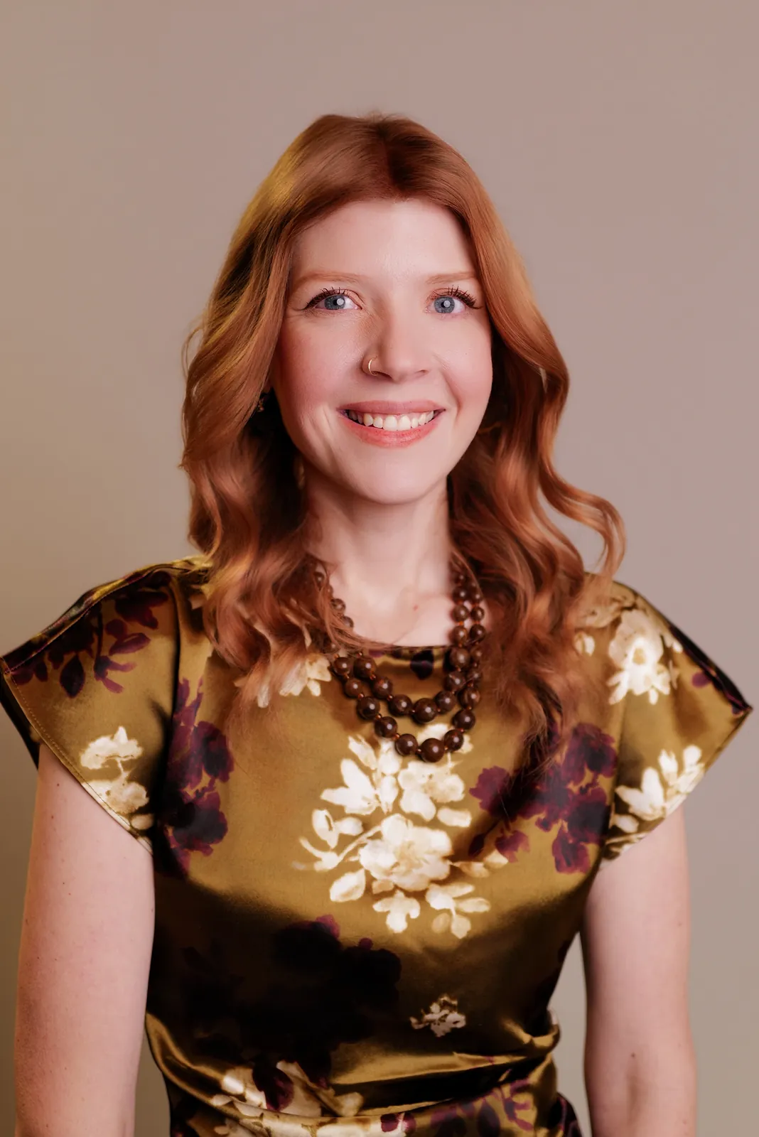 Studio headshot of a smiling woman with auburn hair in a floral print dress on a beige backdrop