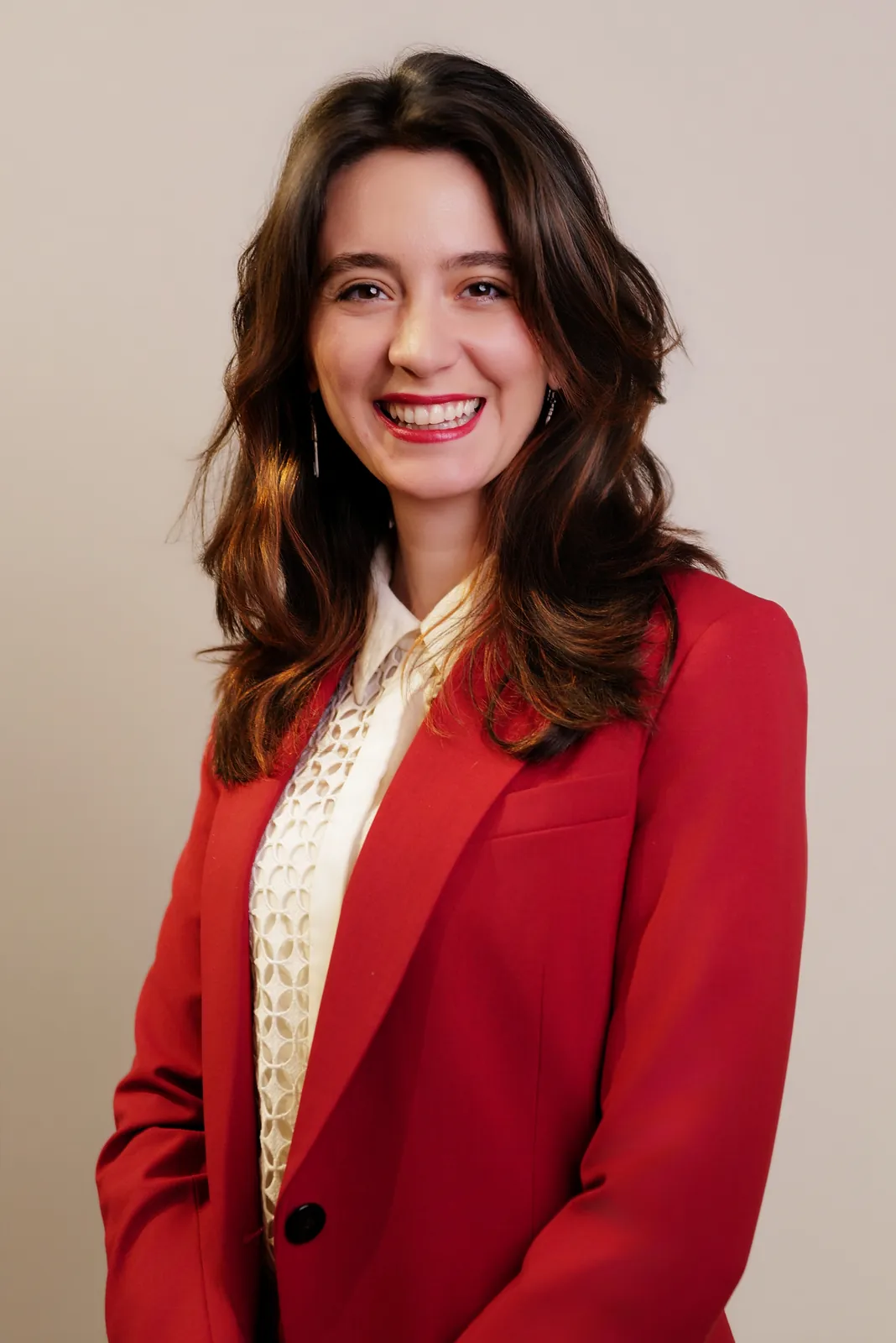 Studio headshot of a smiling brunette woman in a red blazer on a beige backdrop
